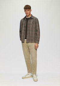 Karierte Flanellbluse in Braun- und Grautönen, über einem dunklen Pullover getragen, kombiniert mit beigen Hosen und weißen Turnschuhen. Vereinfachtes, modernes Design.