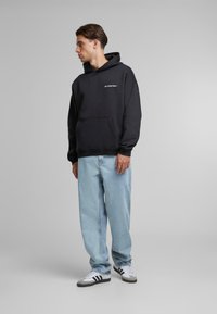 Black hooded sweatshirt with front pocket, white text logo. Worn with light blue loose-fitting jeans and black and white sneakers.