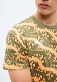 Mies, jolla on päällään vihreän ja oranssin abstraktin kuosin lyhythihainen t-paita, jossa on valkoinen Puma-logo rinnassa.