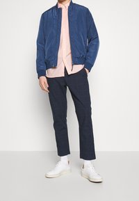 Giacca bomber blu navy, camicia button-up rosa chiaro, pantaloni a quadri blu navy e sneaker bianche. Tessuto morbido, taglio sartoriale e dettagli con zip.