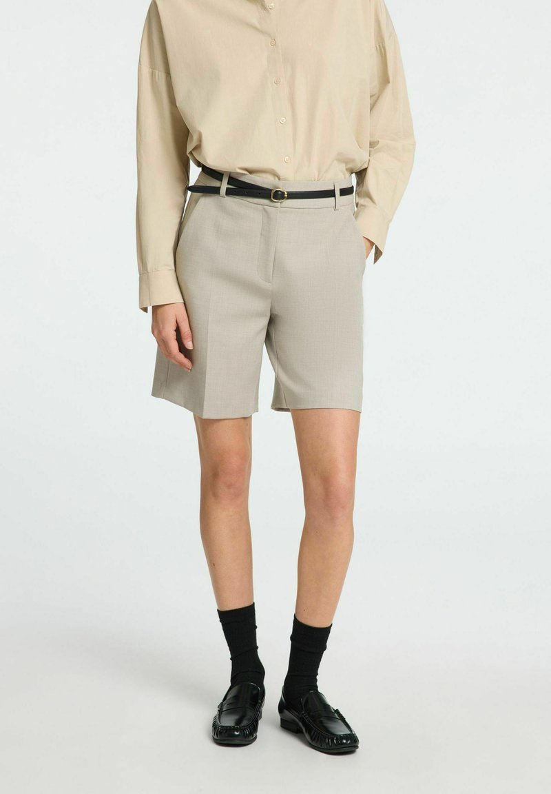 Beige skræddersyede shorts med en glat tekstur, der har en mellemhøj talje, sidelommer og er stylet med et sort bælte og loafers.