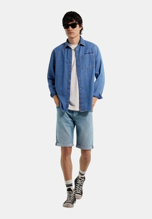 Giovane uomo con occhiali da sole, camicia di jeans blu sopra una t-shirt bianca, pantaloncini di jeans chiari, calzini a righe e sneakers alte nere.