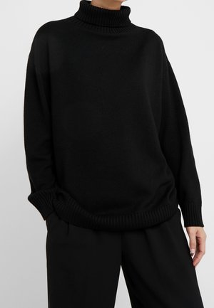 Osoba ubrana w luźny czarny golf i czarne spodnie, stojąca z jedną ręką w kieszeni na tle jasnego, jednolitego tła.