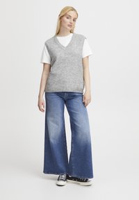 Graham gestrickte Weste mit V-Ausschnitt, getragen über einem weißen T-Shirt. Kombiniert mit weit geschnittenen blauen Jeans und schwarzen Canvas-Sneakern.