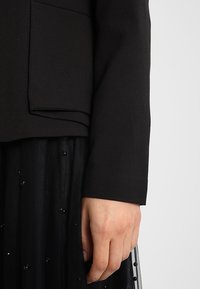 Czarna marynarka o dopasowanym kroju z okrągłą kieszenią, pokazana obok teksturowanej czarnej spódnicy z subtelnymi ozdobami z koralików.