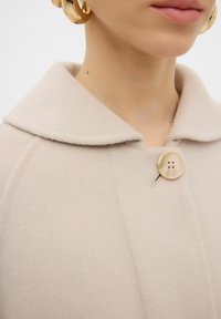 Cappotto in misto lana beige con colletto arrotondato, dotato di un grande bottone e una texture morbida. Design semplice con dettagli di cucitura in evidenza.