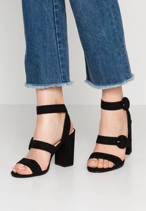 Pieds portant des sandales à talons noirs avec deux brides de cheville et boucles, associés à un jean bleu court avec un ourlet effiloché sur un fond clair.