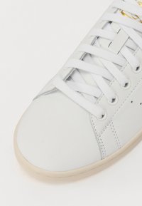 Zapatilla blanca de cuero con una textura suave, que presenta cordones blancos, una punta redondeada y una suela de goma beige. Se puede ver el logo de la marca en dorado.
