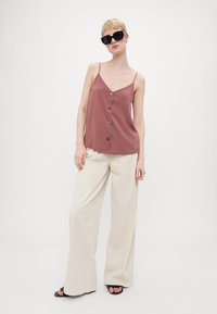 Top sin mangas de color óxido con un escote en V profundo y cinco botones decorativos, combinado con pantalones de pierna ancha en crema. Textura de tela suave.