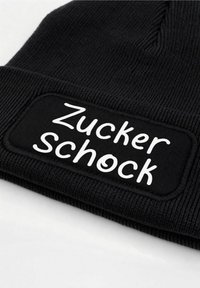 Black knit beanie featuring a rectangular patch with the text "Zucker schock" in white. Ribbed texture and soft material.