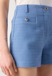 Pantalones cortos de color azul claro con textura y patrón tejido, que cuentan con dos bolsillos frontales y un acento de hardware plateado. Tela suave con un corte entallado.