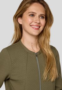 Olive green zip-up jacket with a textured fabric, round neckline, and silver zipper detail, featuring a close-fitting silhouette.