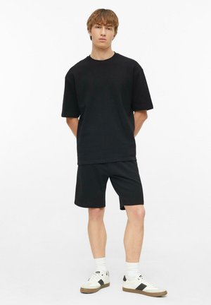 Młody mężczyzna stoi, ubrany w czarny oversize'owy t-shirt i szorty, z białymi skarpetkami i białymi trampkami na jednolitym białym tle.