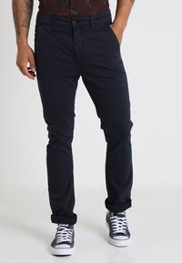 Tummansinisiä puuvillahousuja, joissa on slim fit -malli, napitus, vyölenkit ja käännetyt lahkeet, yhdistettynä valkoisiin lenkkareihin.