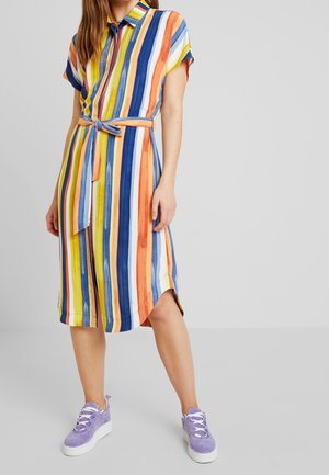 Vrouw draagt een knielange jurk met knopen en verticale strepen in blauw, oranje, geel en wit, gestrikt in de taille, met paarse sneakers.
