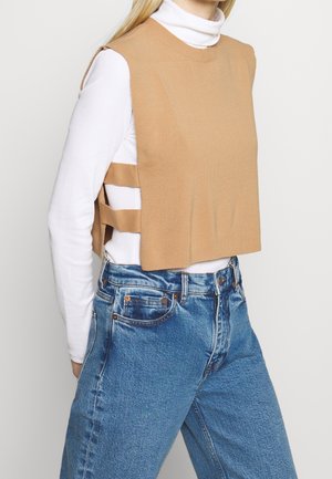 Frau trägt blaue Jeans, weißen Rollkragenpullover und ärmellose, camelfarbene Strickweste mit seitlichen Ausschnitten und horizontalen Riemen.