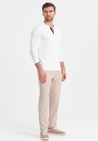Weißes Langarm-Henley-Shirt mit schwarzem Knopfverschluss, kombiniert mit hellbeigen Chinos und beigen Sneakers, minimalistisches Design, glatte Textur.