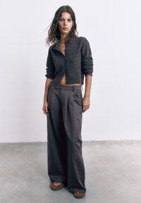 Ung kvinde med bølget hår iført en mørk cropped cardigan, vide bukser og platformsandaler står på en grå gulv mod en hvid væg.