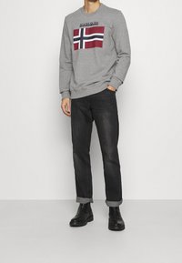 Hombre vestido con un sudadera gris que tiene un gráfico de cruz en rojo, blanco y azul, jeans negros con puños y botas de cuero negras, contra un fondo blanco.
