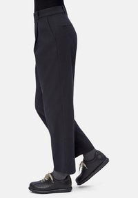 Persona vestida con pantalones negros a medida, calcetines grises y zapatos de cuero negros con cordones, de pie contra un fondo claro. 