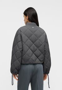 Γκρι μπουφάν με επένδυση καπιτονέ, ψηλό γιακά, ελαστικά κορδόνια στη ραφή και διακριτικό λογότυπο στην πλάτη.