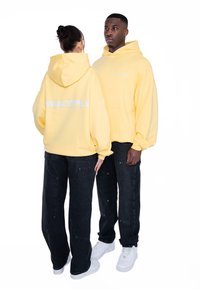 Sudaderas con capucha de suave tela amarilla, con un bolsillo frontal y el logo impreso "MULTIPLY" en la parte posterior. Llevadas con jeans negros holgados.