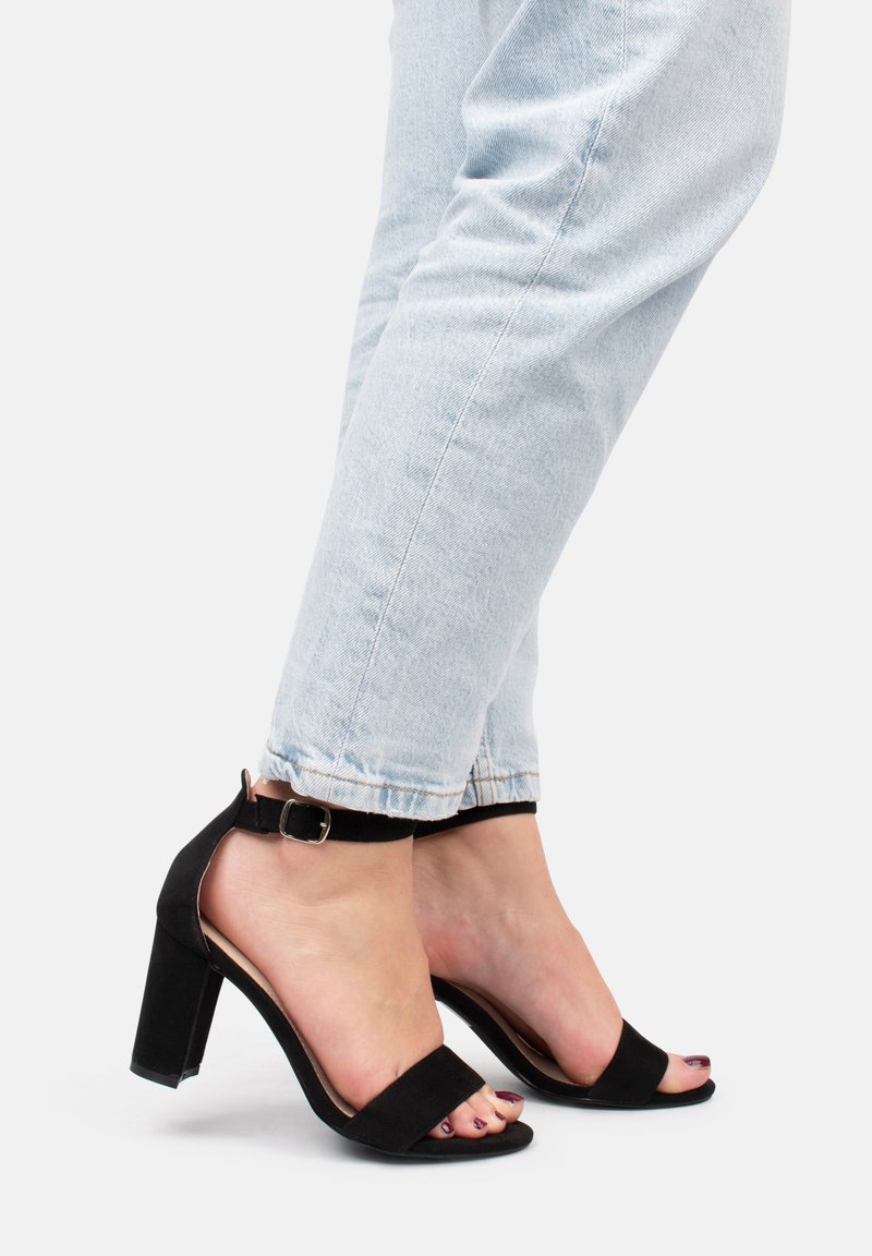 Sandalias de tacón alto de ante negro con tira en el tobillo y diseño de punta abierta. Combinadas con unos jeans de denim azul claro, mostrando un aspecto casual.