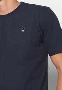 Granatowa bawełniana koszulka z okrągłym dekoltem, krótkimi rękawami i małym okrągłym logo z białą literą "a" na piersi.