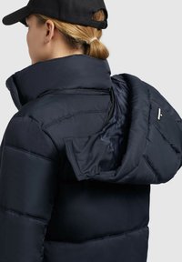 Navy puffer jacket featuring a high collar, partially unzipped hood, and quilted texture. Material appears soft and insulated with metallic accents.