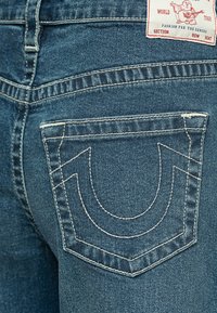 Plave traperice s karakterističnim dizajnom stražnjeg džepa i bijelim šavovima. Materijal ima teksturiranu površinu i moderni kroj.