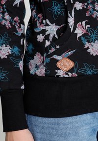 Zwarte sweater met bloemenprint in roze en turquoise, geribde zoom en manchetten, en een bruin leren label met geperst ontwerpdetail.
