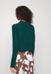 Vrouw met krullend haar in een donkergroene ribgebreide trui en bruine en witte gemusterde broek, met de rug naar de camera tegen een effen achtergrond.
