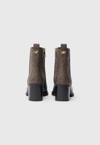 Stivaletti alla caviglia in pelle marrone con suola in pelle nera e tacco a blocco, dotati di chiusura con zip e accentuazione del logo dorato sul retro.