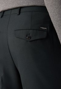 Pantalons noirs en tissu texturé, avec une poche arrière boutonnée et un patch logo. La ceinture est visible en gris.