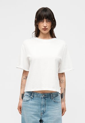 Kobieta o ciemnych włosach nosząca kolczyki koła, prostą białą krótką koszulkę typu cropped z krótkim rękawem oraz jasnoniebieskie dżinsy, pokazując tatuaże na obu przedramionach.