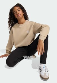 Béžová mikina s okrúhlym výstrihom a bielym logom; čierne nohavice s rebrovanou textúrou a priliehavým strihom; biele a sivé tenisky s čiernymi pruhmi.