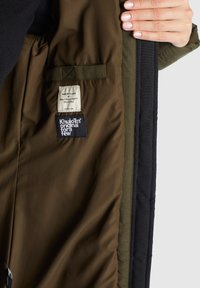 Brun och grön inredning av en jacka, med etiketter med text. Materialet verkar slätt och vattenavvisande.