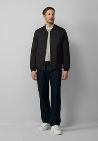 Veste bomber noire avec fermeture éclair à l'avant, poignets et col côtelés ; portée sur un léger pull et un jean foncé ; accompagnée de baskets blanches.