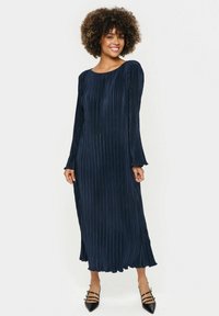 Rochie plisată de culoare bleumarin, cu mâneci lungi, decolteu rotund și fustă fluidă. Prezintă un material texturat și o lungime până la jumătatea gambei. Pantofi negri cu toc.