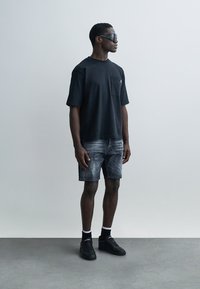 T-shirt en coton noir à manches courtes avec une poche, associé à un short en denim gris et des baskets noires, sur un fond neutre.