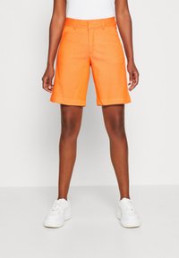 Oranžové lněné šortky s plochým předním dílem, poutky na opasek a zahnoutými lemy, v kombinaci s bílými teniskami, na jednobarevném pozadí.