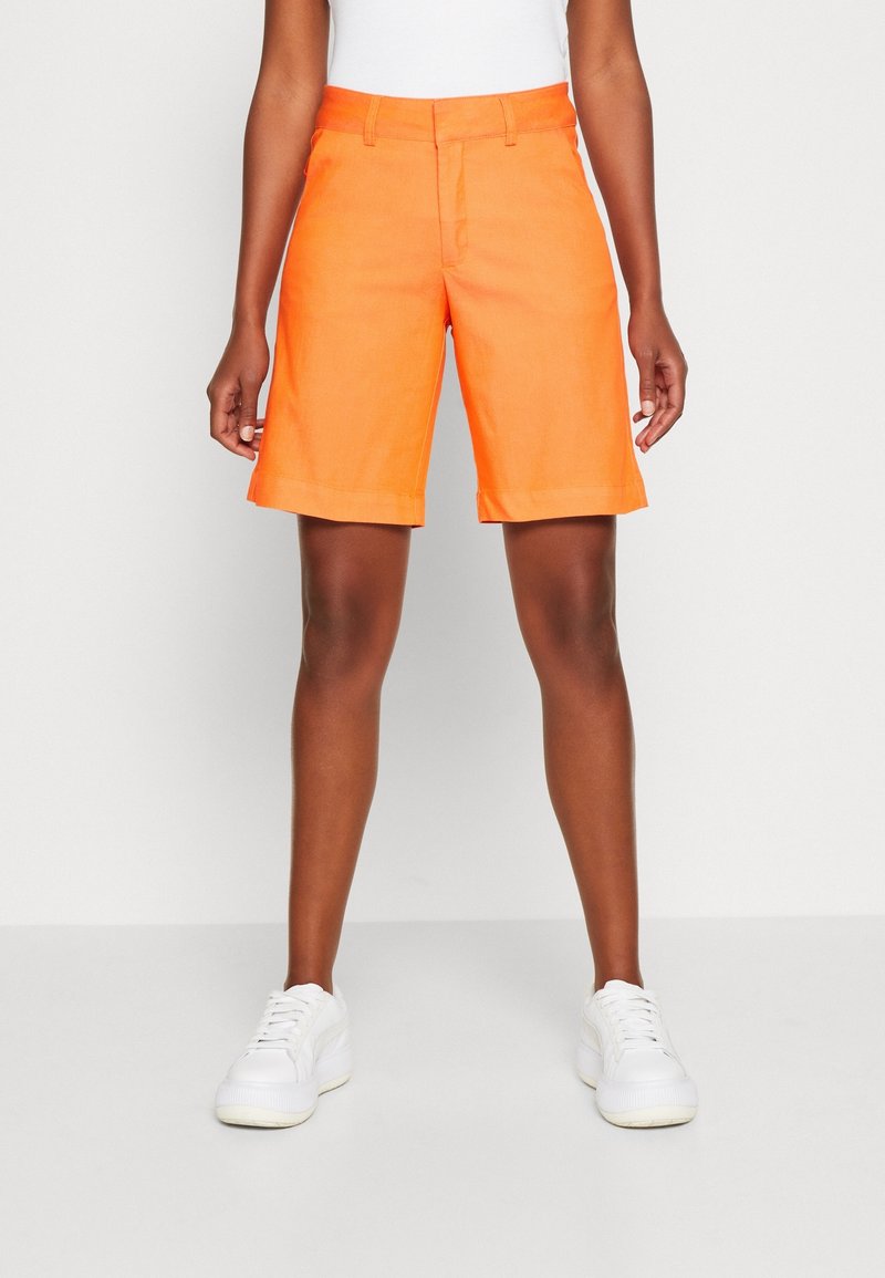 Oranssi pellavashortsit, joissa on litteä etuosa, vyölenkit ja käänteet, yhdistettynä valkoisiin tennareihin, yksinkertaista taustaa vasten.