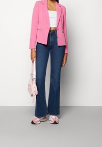 Kobieta w różowej marynarce, białym topie crop, niebieskich dżinsach flare, różowo-białych sneakersach, trzymająca małą różową torebkę na tle jednolitym.