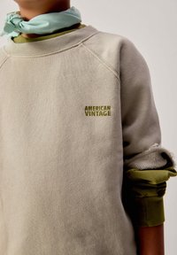 Personne portant un sweat-shirt beige avec le logo "American Vintage" et les manches retroussées, par-dessus une chemise à manches longues verte et un foulard vert clair autour du cou.