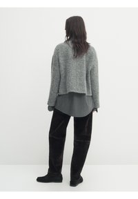 Pull en tricot gris court sur une chemise à longues manches gris et blanche à rayures, associé à un pantalon large en velours marron foncé et des chaussures noires.