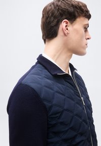 Νεαρός άνδρας σε προφίλ, φορώντας ναυτικό μπουφάν με ραμμένα ανάγλυφα σχέδια και μανίκια από πλεκτό ύφασμα, με ασημένιο φερμουάρ, μπροστά σε απλό φωτεινό φόντο.