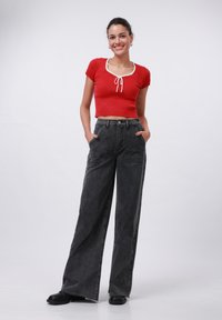 Top court rouge à côtes avec bordure blanche et cordon de serrage ; pantalon large en denim noir, ample ; modèle portant des chaussures noires, les mains dans les poches.