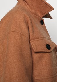 Nærbillede af en brun uldjakke med rejst krave og en knaplukket klaplomme på brystet.