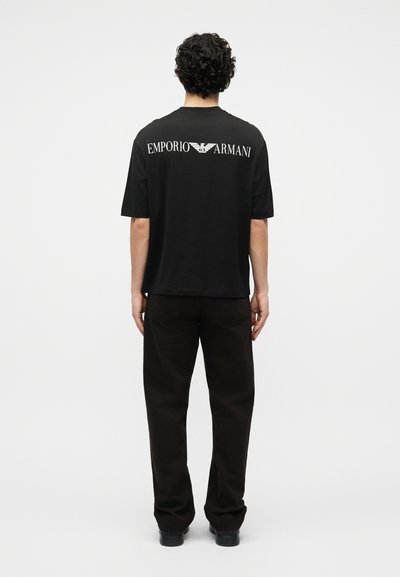 Άνδρας με σγουρά μαλλιά, φοράει μαύρο t-shirt Emporio Armani και μαύρο παντελόνι, στέκεται με την πλάτη σε ένα απλό φωτεινό φόντο.