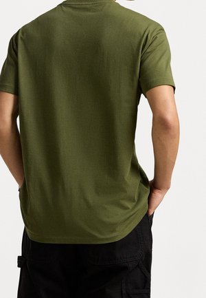 Mann trägt ein olivgrünes T-Shirt mit kurzen Ärmeln und Händen in den Taschen, von hinten vor einem einfarbigen Hintergrund gezeigt.
